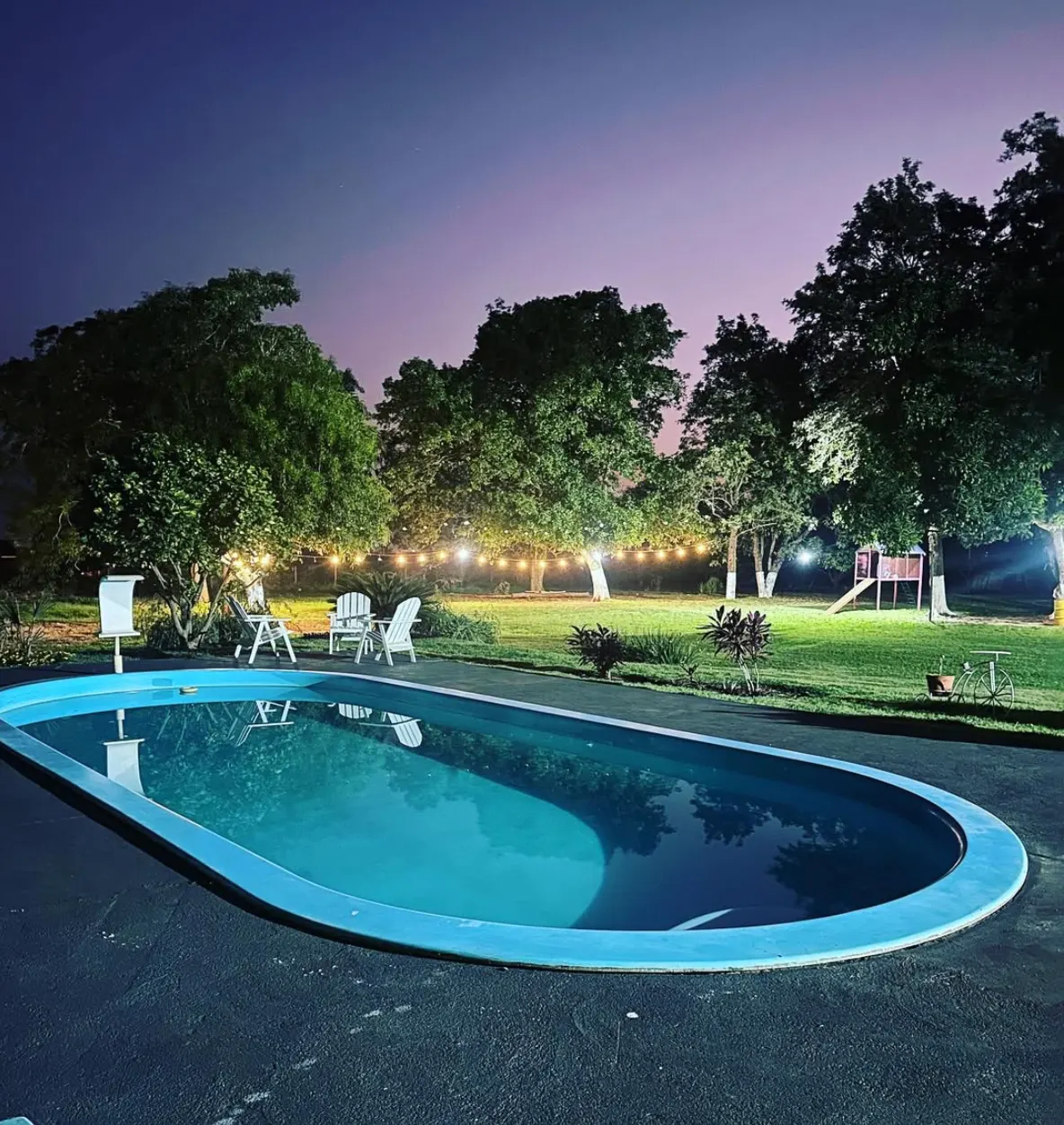 Piscina oval iluminada à noite, com varal de luzes aceso entre as árvores e céu arroxeado